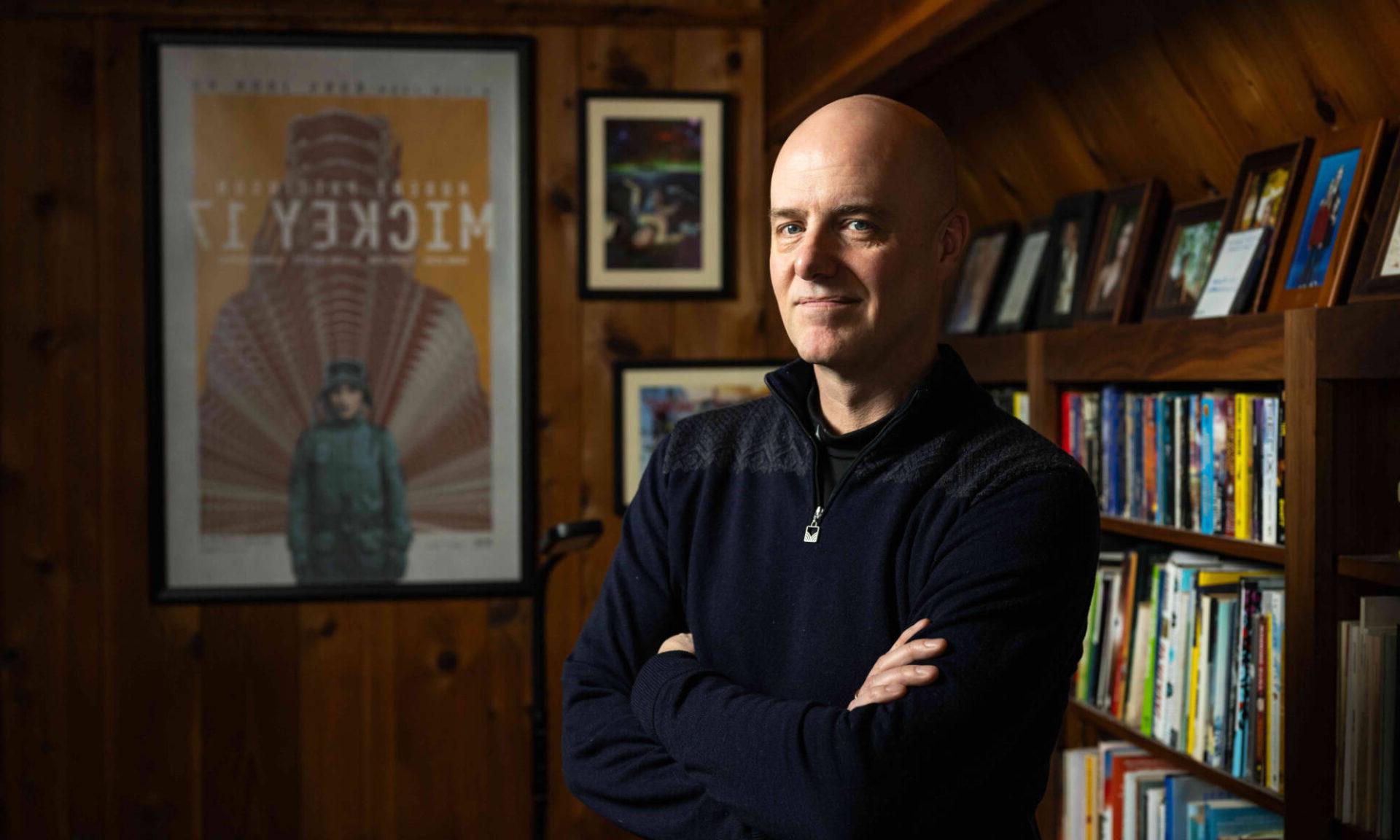 Edward Ashton, author of Mickey7, looks at the camera with his arms folded. Behind him is an extensive bookshelf and a framed movie poster for Mickey7.